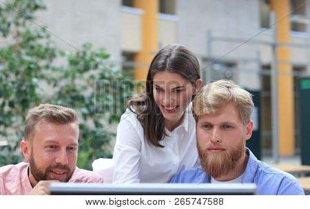 Group Of Young People In Casual Wear Sitting At The Office Desk And Discussing Something While Looki