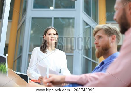 Group Of Young People In Casual Wear Sitting At The Office Desk And Discussing Something While Looki