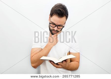 Thoughtful Young Man In Glasses Standing And Reading Book. He Holds It In Left Hand. Another One In 