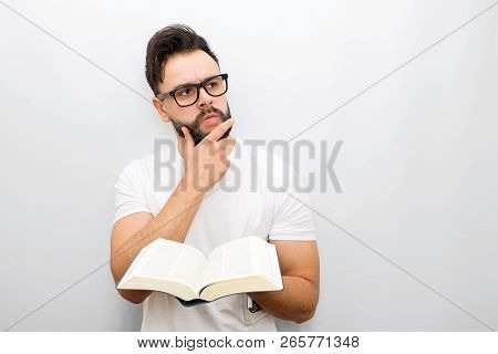 Wize And Thoughtful Young Man In Glasses Stand And Hold Opened Book In Hands. He Looks To Side. Guy 