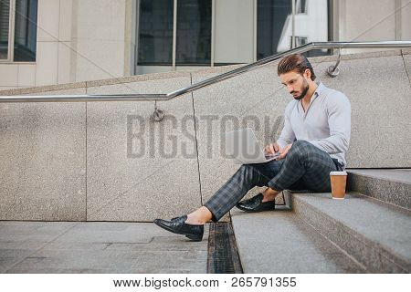 Calm And Peaceful Young Man Sits On Steps And Works. He Looks At Laptop Screen. There Are Plastic Cu
