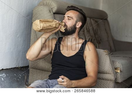 Drunkard Sitting On A Building Rooftop Terrace, Holding A Bottle In A Paper Bag, Drinking