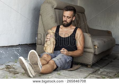 Drunkard Sitting On A Building Rooftop Terrace, Holding A Bottle In A Paper Bag, Drinking