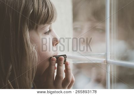 Sad Caucasian Girl Standing By The Window And Playing Harmonica At Home