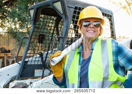 Smiling Female Worker Holding Technical Blueprints Near Small Bulldozer At Constrcution Site
