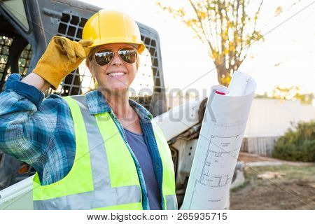 Smiling Female Worker Holding Technical Blueprints Near Small Bulldozer At Constrcution Site