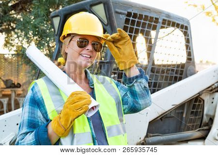 Smiling Female Worker Holding Technical Blueprints Near Small Bulldozer At Constrcution Site