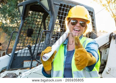 Smiling Female Worker With Thumbs Up and Technical Blueprints Near Small Bulldozer At Constrcution Site