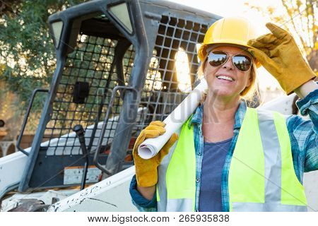 Smiling Female Worker Holding Technical Blueprints Near Small Bulldozer At Constrcution Site