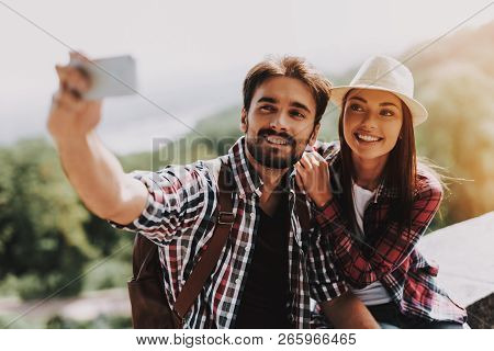 Couple Sitting On Concrete Ledge Taking Selfie. Young Man And Attractive Woman With Backpacks Sittin