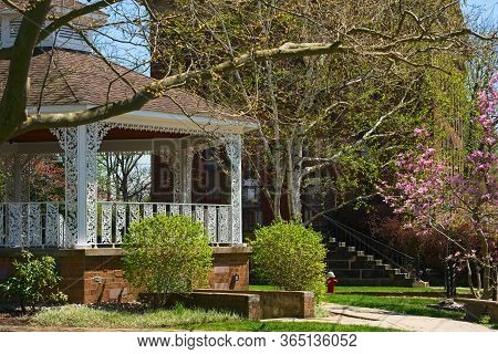 A Gazebo With Fancy Decorative Grillwork Painted White Stands On A Village Square In Spring With A M