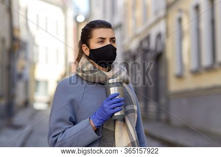 health, safety and pandemic concept - young woman wearing black face protective reusable barrier mask with tumbler or thermo cup on city street