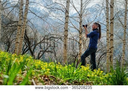 A Man Photographer Stands On The Trail In The Spring Birch Forest. The Guy Holds A Camera In His Han