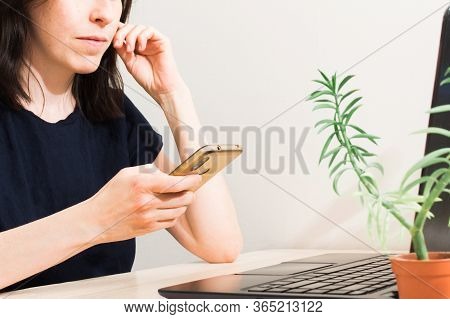 Beautiful Young Woman With Dark Hair Sitting At Table With Laptop And Using Phone. Online Business W