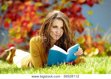 Jeune femme se trouve sur l'herbe verte et lit livre contre automne parc.