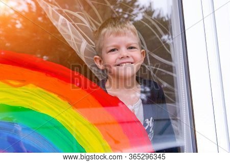 Little Blonde Boy With Rainbow On Window During Covid-19 Quarantine At Home. Stay At Home Social Med