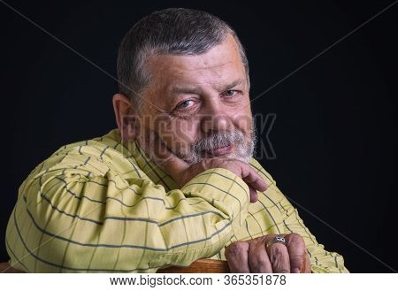 Nice Portrait Of A Smiling Senior Man In Yellow Shirt Leant Elbow On  A Back Of Chair Against Black 