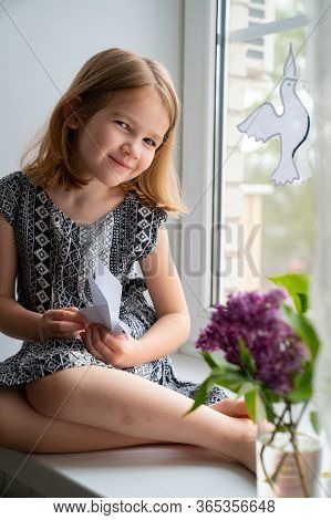 Little Girl Kid Sitting On The Windowsill With Origami In His Hands And On The Window Of Doves Symbo