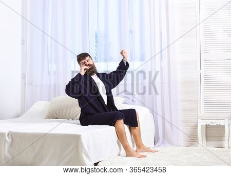 Guy On Sleepy Face Yawning And Stretching. Awakening Concept. Man In Robe Sits On Bed, White Curtain