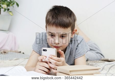 A Boy With A Smartphone In His Hands Next To Book In Bed Teaches Lessons. Distance Learning Online O
