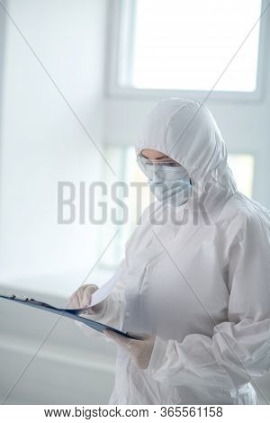 Medical Worker In Protective Clothing, Medical Mask And Goggles Looking Through Papers On Clip Folde