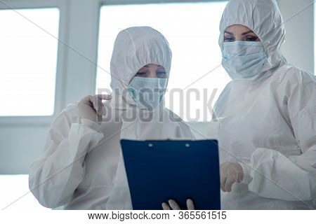Medical Workers In Protective Clothing And Medical Masks Reading Something On Clip Folder