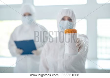 Medical Worker In Protective Clothing Showing Disinfecting Wipes Bottle, Her Colleague Standing Behi