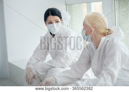 Two Female Medical Workers In Protective Clothing And Medical Mask Sitting Next To The Window, Discu
