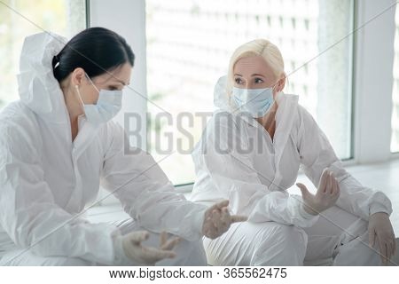 Two Female Medical Workers In Protective Clothing And Medical Mask Sitting Next To The Window, Talki