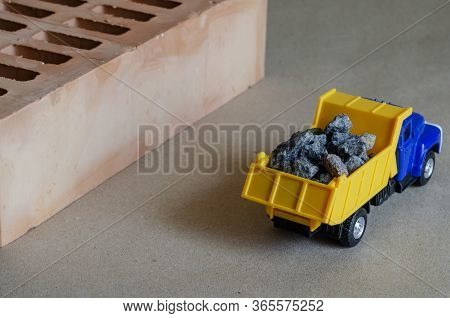 A Yellow Dump Truck Carries Rubble Next To A Brick. Toy Truck With A Full Body Of Building Materials