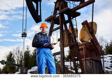 Portrait Of Petrolium Engineer In Blue Overalls And Orange Helmet, With Laptop, Standing With His Ba