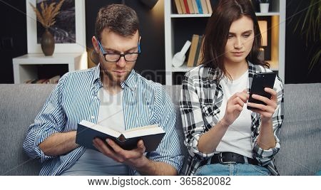 Two People Trying To Divert Themselves Sitting On Couch, Woman Surfing Cellphone While Man Reading B