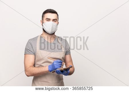 Young seious waiter in apron, protective mask and gloves writing down your order in notepad while working in cafe or restaurant