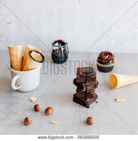 Homemade Dark Chocolate Brownies, Sauce In Glass Jar, Ice Cream Waffle Cones And Cupcake On Gray Bac
