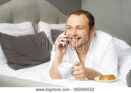 A Man In A Bathrobe Is Lying In A White Bed Drinking Coffee And Talking On The Phone. Muzhchina On A