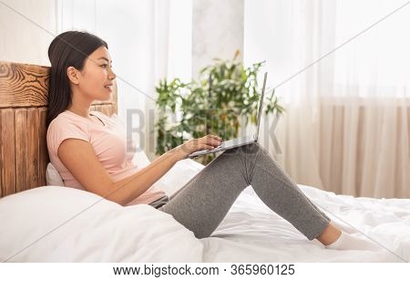 Homey Asian Woman Using Laptop Sitting In Bed At Home Browsing Internet In The Morning. Weekend Leis