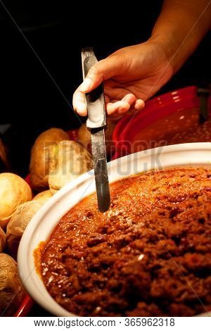 Bowl Of Chilli Being Stirred With A Black Handles Spoon With Jacket Potatoes Next To The Bowl,