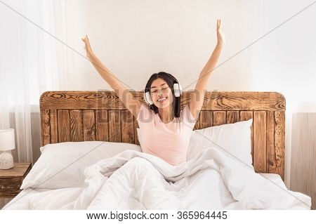 Happy Asian Girl Listening To Music In Wireless Headphones Sitting In Bed Waking Up In The Morning A
