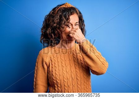 Middle age beautiful woman wearing casual yellow sweater over isolated yellow background smelling something stinky and disgusting, intolerable smell, holding breath with fingers on nose. Bad smell