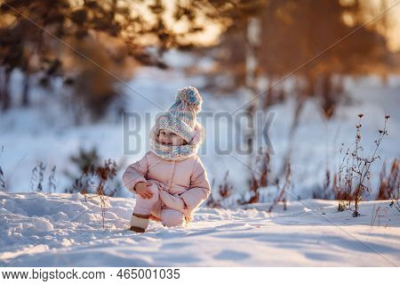 An Adorable Little Girl In A Warm Coat And A Colorful Hat With A Pom-pom Stands In The Snow On The S