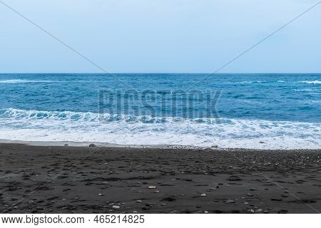Black Sand On The Beach Against Blue Turbulent Water With White Foam. Empty Volcanic Sand Beach On T