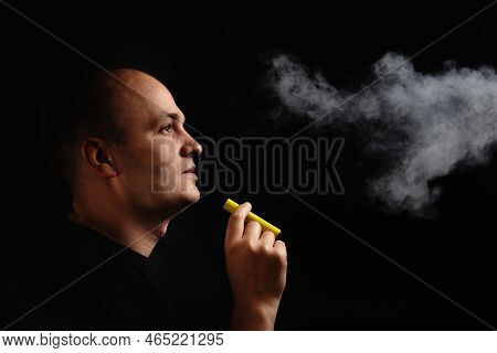 A Young Man Smokes An Electronic Cigarette Against A Black Background And Blows Smoke. E-cigarette S