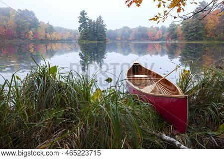 Red Wood Canoe On Shore Of A Small Lake With An Island On A Cloudy Autumn Morning