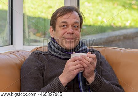 An Elderly Man Of 45-50 Years Old In A Scarf Holds A White Mug With Both Hands While Sitting On The 