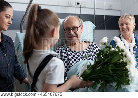 Sick Grandfather Receiving Flowers From Granddaughter In Hospital Room. Bedridden Old Man Being Visi