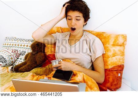 Teenager Sitting In Bed Under Blanket, Holding Coffee Cup, Working Or Studying Online On Laptop With