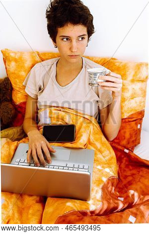 Teenager Sitting In Bed Under Blanket, Holding Coffee Cup, Working Or Studying Online On Laptop With