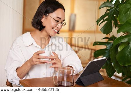 Asian Woman In Glasses, Watching Smth On Digital Tablet, Drinking Coffee In A Cafe And Smiling, Work