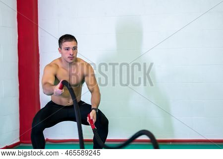 A Handsome Muscular Young Man Using Battle Ropes For Exercise In A Gym. He Focuses On Performing The