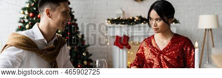 Smiling Man Looking At Serious Asian Wife In Red And Shiny Blouse While Celebrating Christmas At Hom